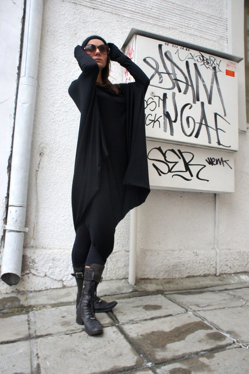 Op de afbeelding: Een vrouw draagt een zwarte lange mouwen tuniek met een cowl hals en zwarte leggings. Ze draagt een zonnebril en zwarte laarzen. De achtergrond is een witte muur met graffiti.