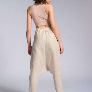 May include: A woman wearing a light brown tank top and beige linen pants with a loose, baggy fit. The pants have a drawstring waistband and a single pocket on the back.