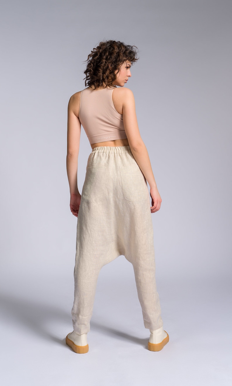 May include: A woman wearing a light brown tank top and beige linen pants with a loose, baggy fit. The pants have a drawstring waistband and a single pocket on the back.