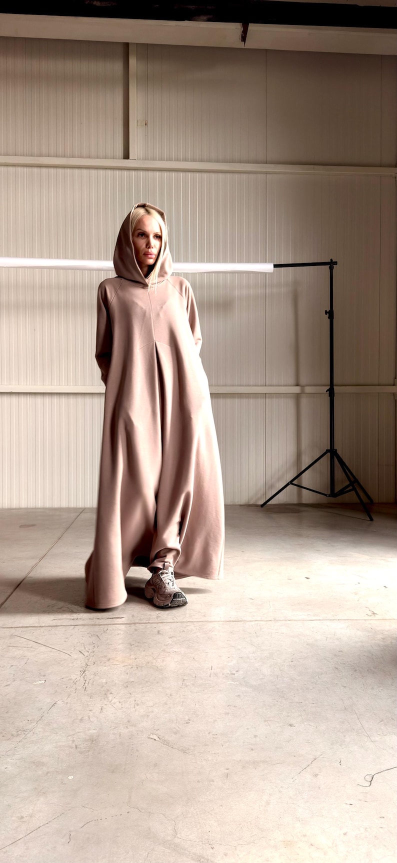 May include: A long, hooded, taupe-colored garment. The garment has a loose, flowing design and is worn with sneakers. The model is standing in a studio setting with a neutral background and a light fixture. Hooded dress from cotton with side pockets and plated front and back