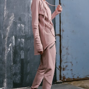 Peut inclure: Une femme portant un sweat &agrave; capuche asym&eacute;trique &agrave; manches longues de couleur rose poussi&eacute;reux avec un cordon de serrage et un pantalon de surv&ecirc;tement assorti. Elle porte des baskets color&eacute;es.