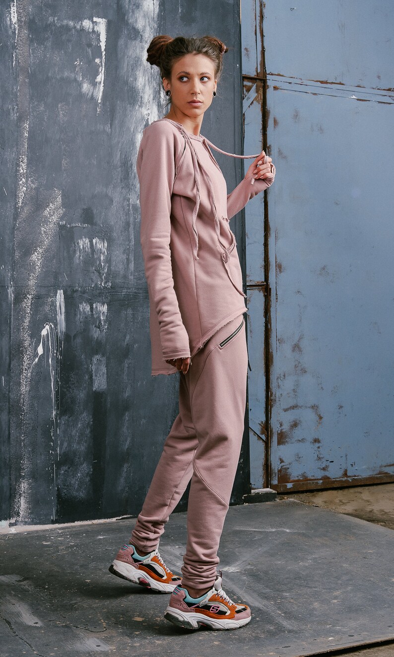 Peut inclure: Une femme portant un sweat &agrave; capuche asym&eacute;trique &agrave; manches longues de couleur rose poussi&eacute;reux avec un cordon de serrage et un pantalon de surv&ecirc;tement assorti. Elle porte des baskets color&eacute;es.