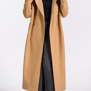 May include: A long camel-colored coat with a double-breasted closure and a high collar. The coat is worn over a black turtleneck and gray pants. The person is wearing black and white high-top sneakers.