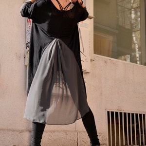 May include: A woman wearing a black sheer tunic with a long, flowing skirt. She is wearing black leggings and black heeled boots. She is standing in front of a window with a white frame.