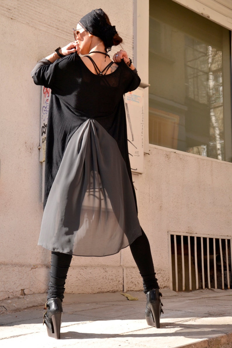 May include: A woman wearing a black sheer tunic with a long, flowing skirt. She is wearing black leggings and black heeled boots. She is standing in front of a window with a white frame.