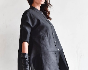 Pode incluir: Uma camisa de linho preta com uma bainha longa e fluida e uma sobreposição transparente. A camisa tem um fecho de botões na frente e uma gola alta. A modelo está usando calças de couro pretas e luvas de couro pretas.