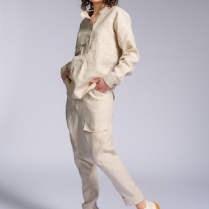 May include: A woman wearing a light beige linen pantsuit with a collared shirt and long sleeves. She is wearing white high-top sneakers with a tan sole.