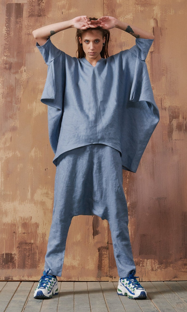 May include: A person wearing a light blue linen jumpsuit with a loose-fitting top and pants. The pants have a wide leg and a gathered detail at the ankles. The person is wearing white sneakers with blue and green accents.