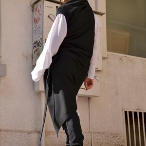 May include: A woman wearing a long black sleeveless top over a white shirt, black pants, and white sneakers with a floral pattern. She is standing in front of a white wall with a window and a metal box.