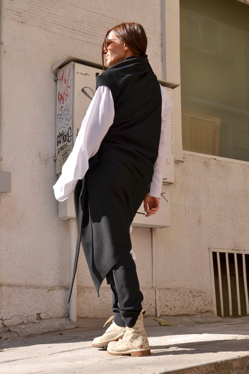 May include: A woman wearing a long black sleeveless top over a white shirt, black pants, and white sneakers with a floral pattern. She is standing in front of a white wall with a window and a metal box.
