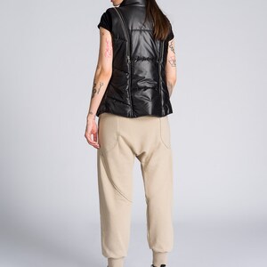May include: A black puffer vest with a high collar and a zip closure. The vest is paired with beige sweatpants and black and white high-top sneakers with a star design. The outfit is set against a plain gray background.