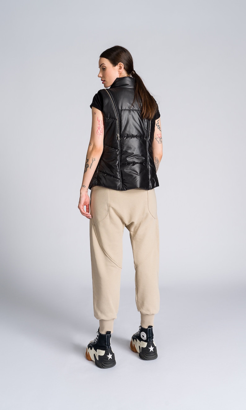 May include: A black puffer vest with a high collar and a zip closure. The vest is paired with beige sweatpants and black and white high-top sneakers with a star design. The outfit is set against a plain gray background.