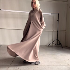 May include: A long, flowing, hooded dress in a muted rose color. The dress features a relaxed fit with a subtle A-line silhouette and pockets. The model is wearing gray sneakers. The fabric appears to be a soft, comfortable material.