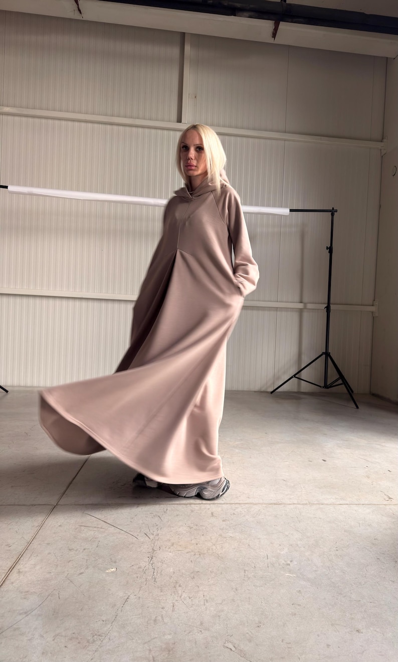 May include: A long, flowing, hooded dress in a muted rose color. The dress features a relaxed fit with a subtle A-line silhouette and pockets. The model is wearing gray sneakers. The fabric appears to be a soft, comfortable material.