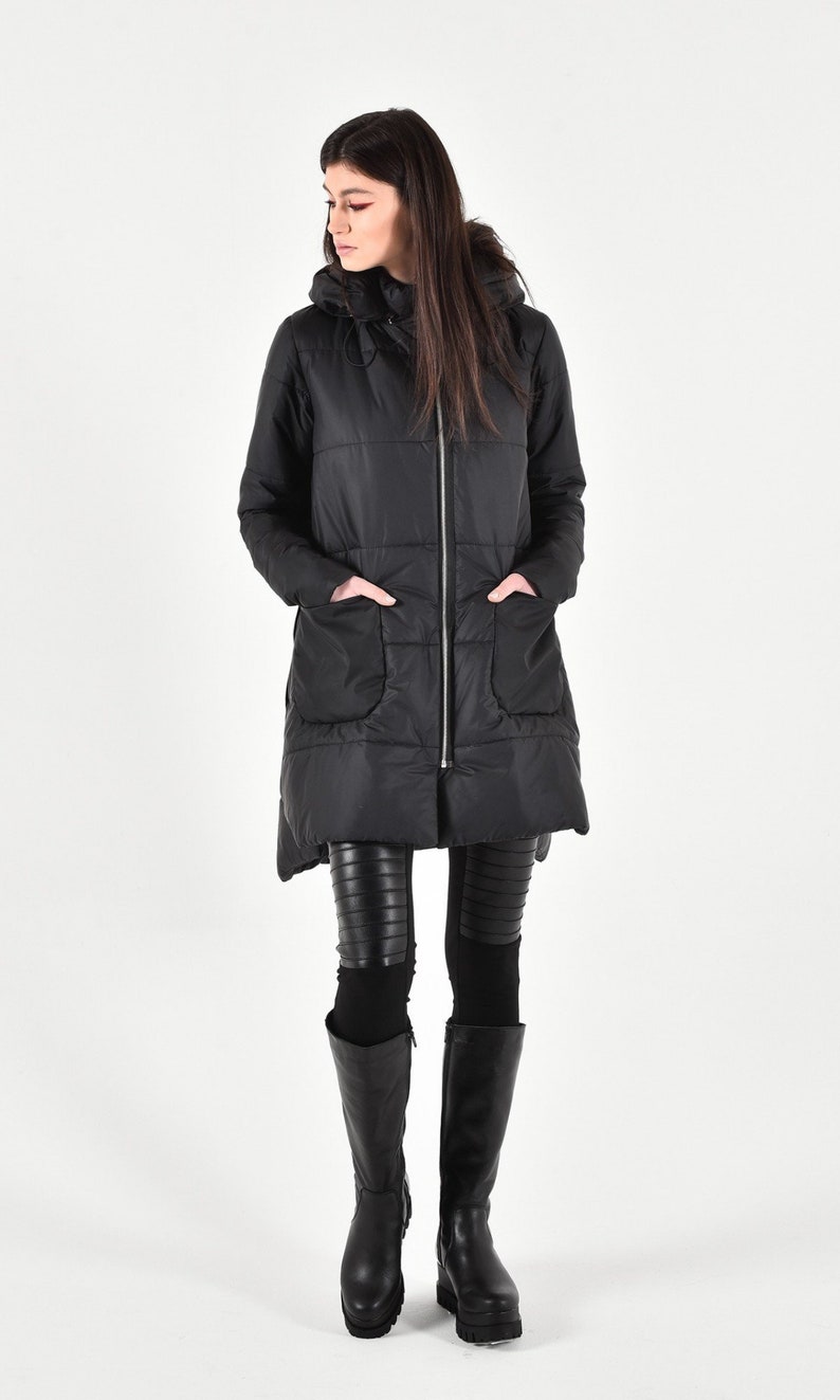 May include: A black long puffer jacket with a hood and zipper closure. The jacket has a quilted design and large pockets. The model is wearing black leggings and tall black boots.