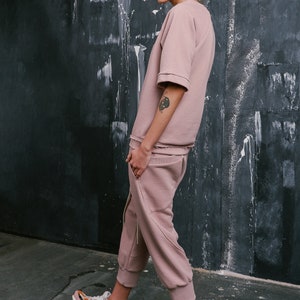 Op de afbeelding: Een vrouw draagt een stoffig roze sweatshirt en een bijpassende joggingbroek. Ze draagt kleurrijke sneakers en heeft haar haar in een knotje.