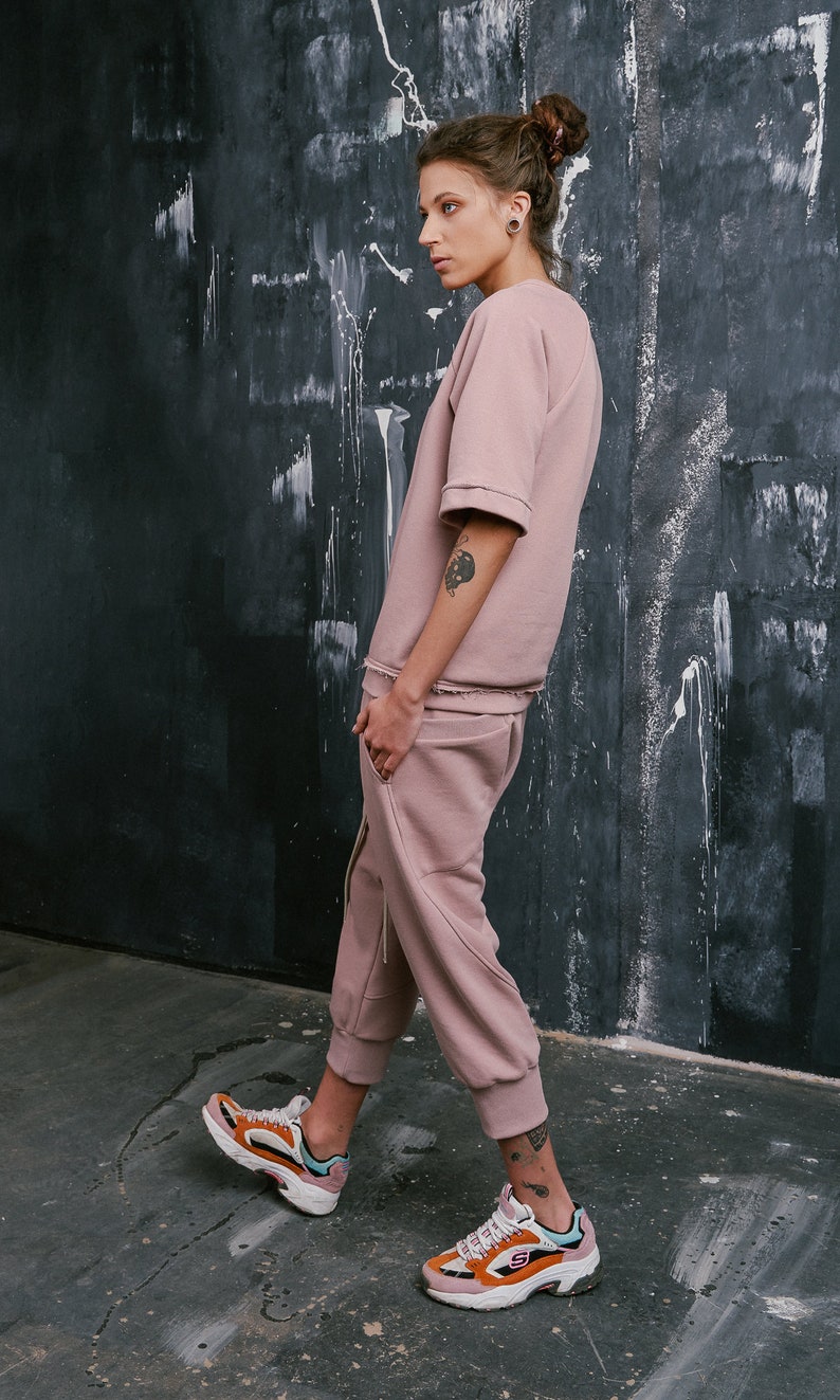 Op de afbeelding: Een vrouw draagt een stoffig roze sweatshirt en een bijpassende joggingbroek. Ze draagt kleurrijke sneakers en heeft haar haar in een knotje.