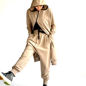 Puede incluir: Una sudadera con capucha beige y pantalones deportivos a juego con cintura elástica. La sudadera tiene cierre de cremallera y los pantalones tienen un diseño largo y fluido.