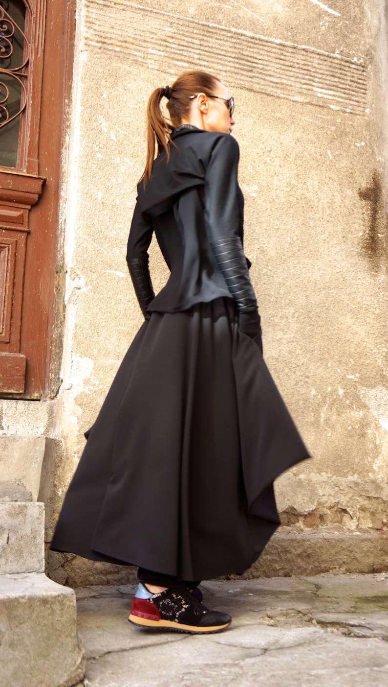 May include: A black hooded jacket with a long, flowing skirt. The jacket has leather accents on the sleeves and the skirt has a unique asymmetrical design. The person is wearing black sneakers with white laces.