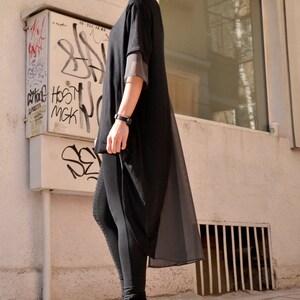May include: A woman wearing a black long-sleeved tunic with a sheer overlay and black leggings. She is wearing black platform shoes with gold buckles. The woman is standing in front of a building with a window and graffiti on the wall.