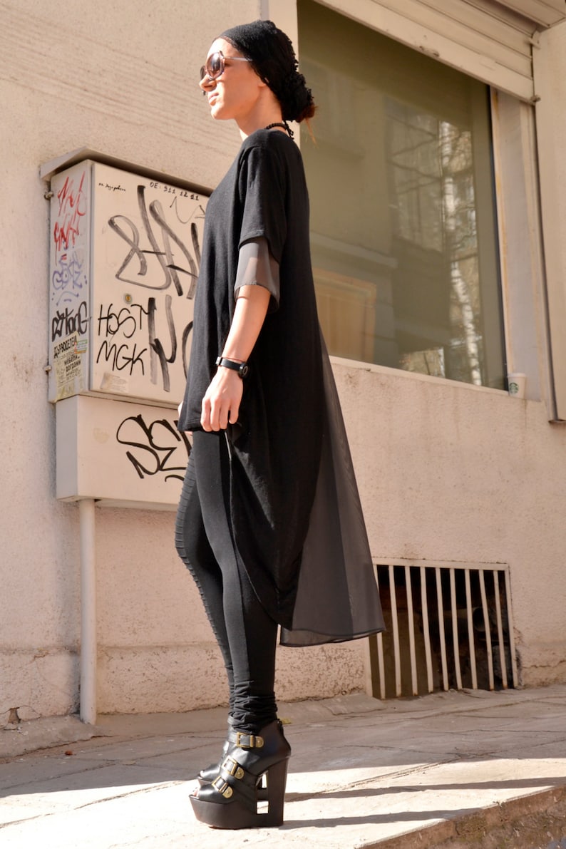May include: A woman wearing a black long-sleeved tunic with a sheer overlay and black leggings. She is wearing black platform shoes with gold buckles. The woman is standing in front of a building with a window and graffiti on the wall.