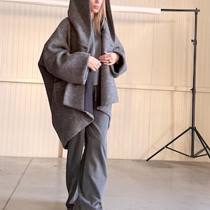 Könnte beinhalten: Ein grauer Kapuzenmantel und eine passende Hose. Der Mantel hat eine große Kapuze und ein lockeres, fließendes Design. Die Hose ist weit geschnitten. Das Outfit besteht aus einem weichen, strukturierten Stoff. Das Model trägt hellfarbene Sneaker.
