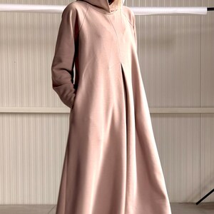 May include: A long, hooded dress in a muted rose color. The dress features a flowing silhouette, long sleeves, and pockets. The model is wearing a black top underneath. The dress is made of a soft, draping fabric.