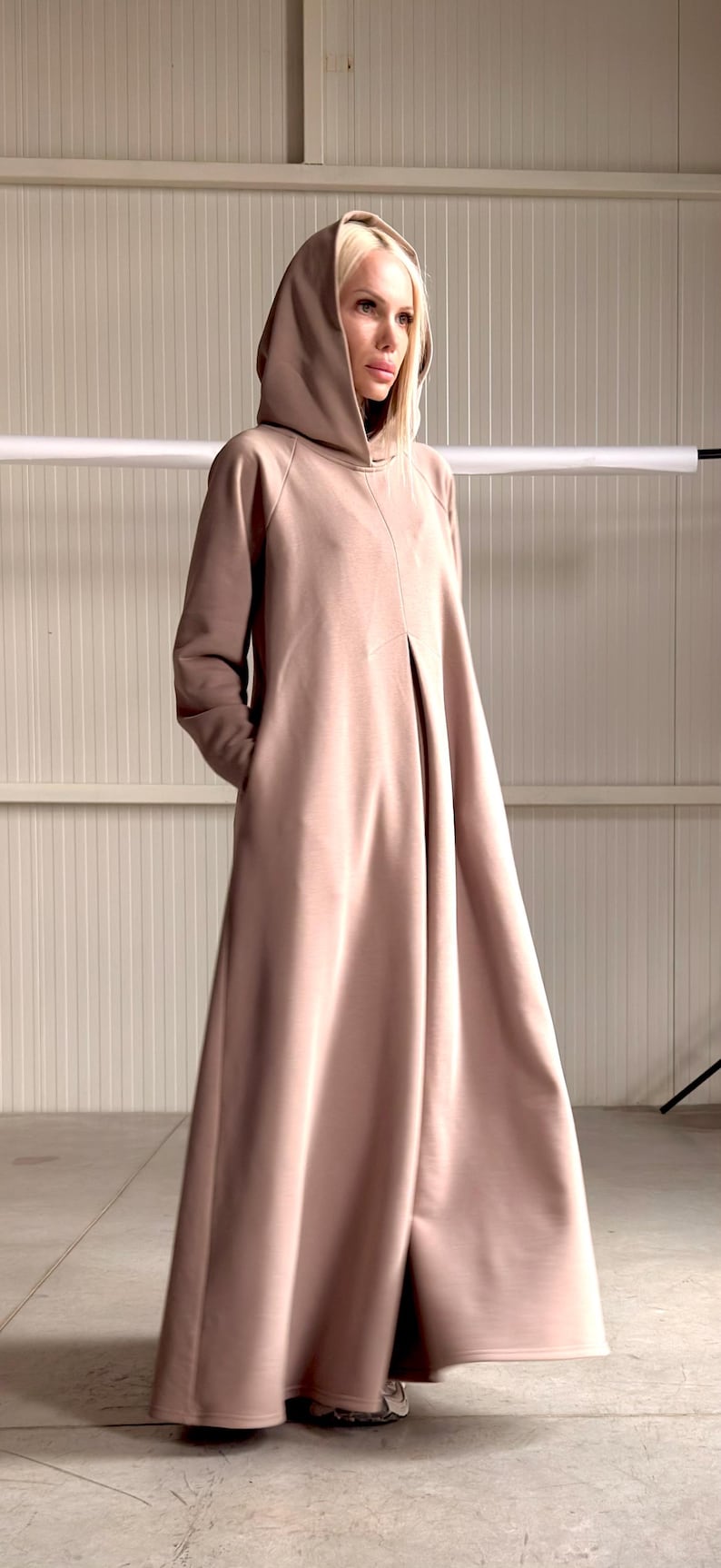May include: A long, hooded dress in a muted rose color. The dress features a flowing silhouette, long sleeves, and pockets. The model is wearing a black top underneath. The dress is made of a soft, draping fabric.