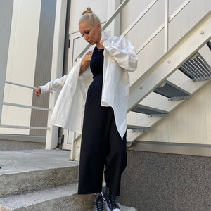 May include: A woman wearing a black tank top, black wide-leg pants, and a white button-down shirt. She is also wearing black and white sneakers with a brown sole. The outfit is styled on a concrete staircase next to a metal staircase.