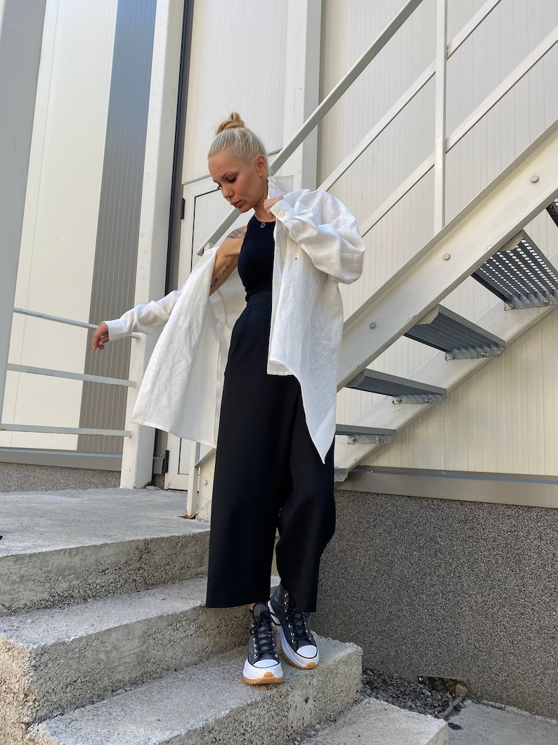 May include: A woman wearing a black tank top, black wide-leg pants, and a white button-down shirt. She is also wearing black and white sneakers with a brown sole. The outfit is styled on a concrete staircase next to a metal staircase.