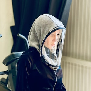 Może przedstawiać: Osoba w szarej bluzie z kapturem z przezroczystym plastikowym osłoną twarzy przymocowaną do kaptura. Bluza ma czarny zamek błyskawiczny, a osłona twarzy jest przezroczysta i ma czarną ramkę.