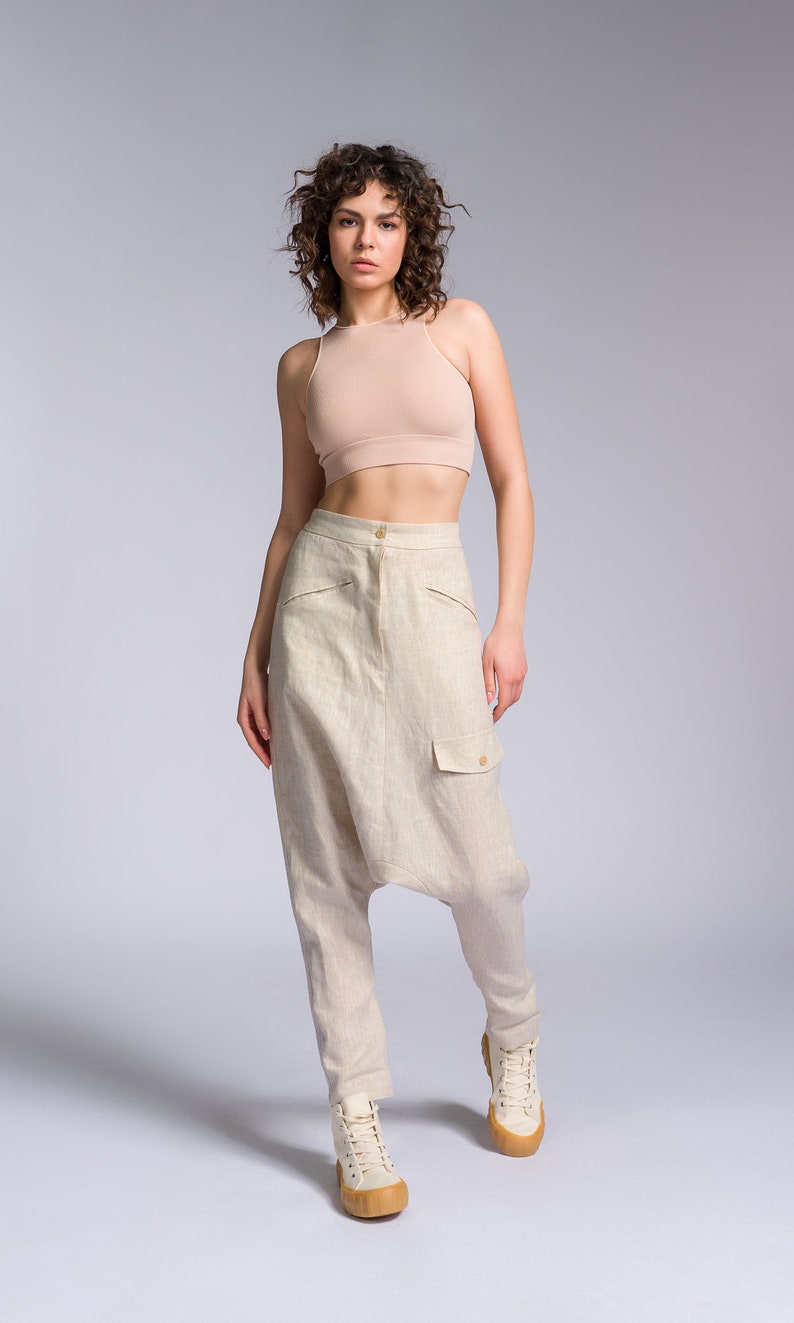 May include: A woman wearing a light brown tank top and beige linen pants with a large cargo pocket. She is wearing white high-top sneakers with a brown sole.