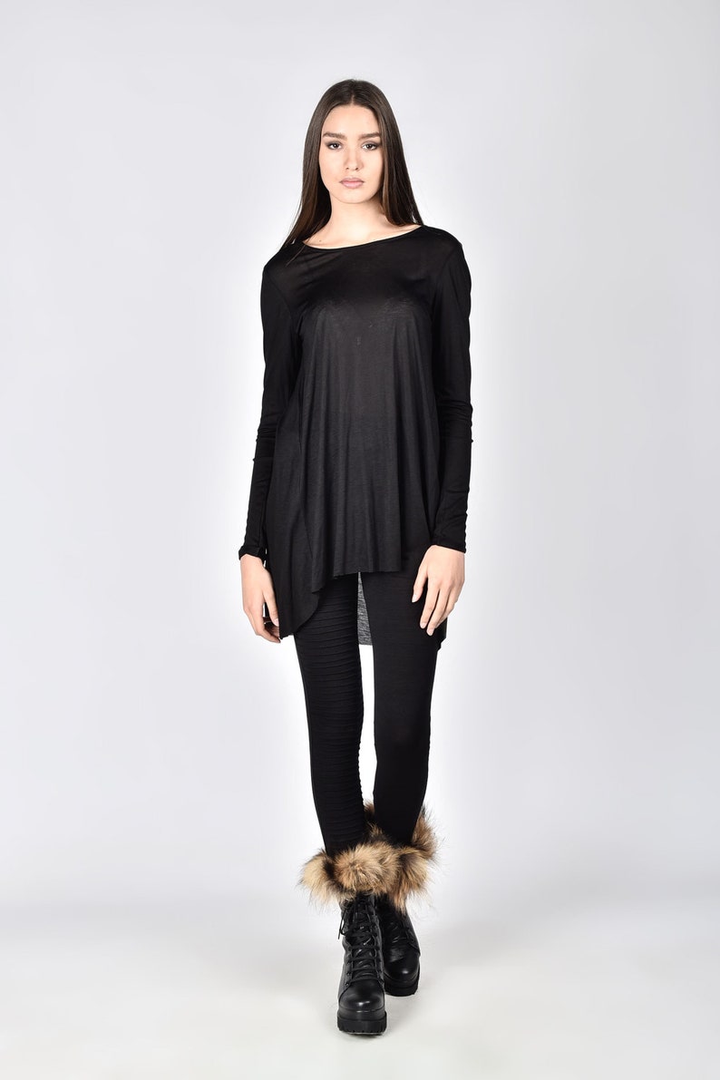 Op de afbeelding: Een persoon draagt een zwarte tuniek met lange mouwen en een zwarte legging. De tuniek heeft een asymmetrische zoom. Ze draagt zwarte laarzen met bontafwerking. De achtergrond is effen wit.