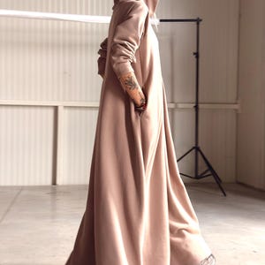 May include: A long, hooded dress in a soft, muted peach color. The dress features a flowing silhouette, a hood, and pockets. The model is wearing sneakers. The overall style is casual and comfortable.