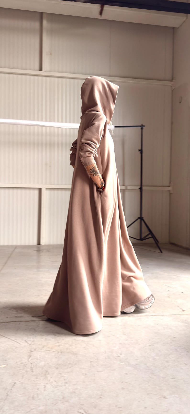 May include: A long, hooded dress in a soft, muted peach color. The dress features a flowing silhouette, a hood, and pockets. The model is wearing sneakers. The overall style is casual and comfortable.