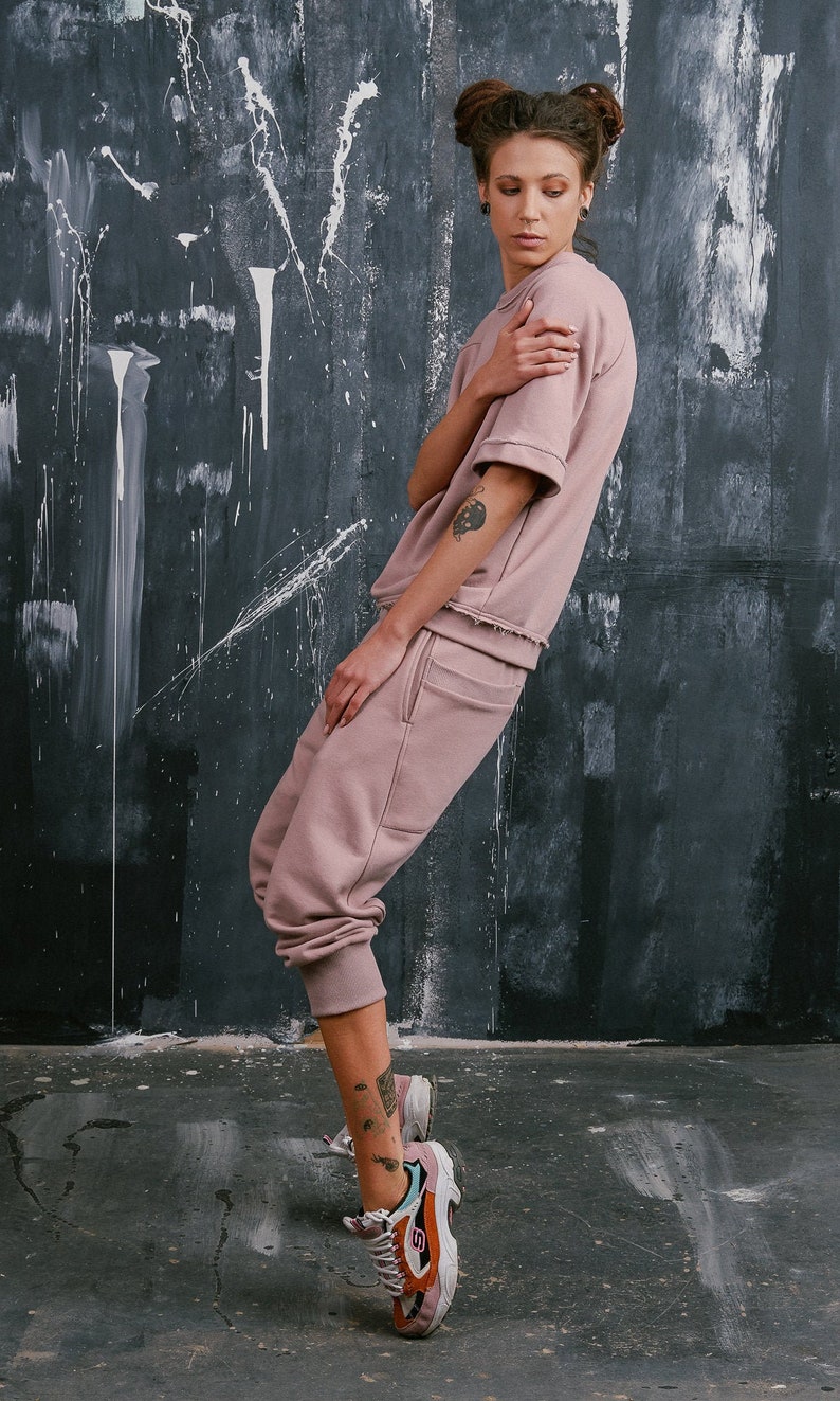 Op de afbeelding: Een vrouw draagt een stoffig roze sweatshirt en een bijpassende joggingbroek. Ze draagt sneakers met oranje en witte accenten. De vrouw heeft haar haar in twee knotjes.