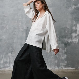 May include: A woman wearing a white linen shirt and black wide-leg pants. She is wearing white sneakers with red accents and black high-top sneakers with red accents.