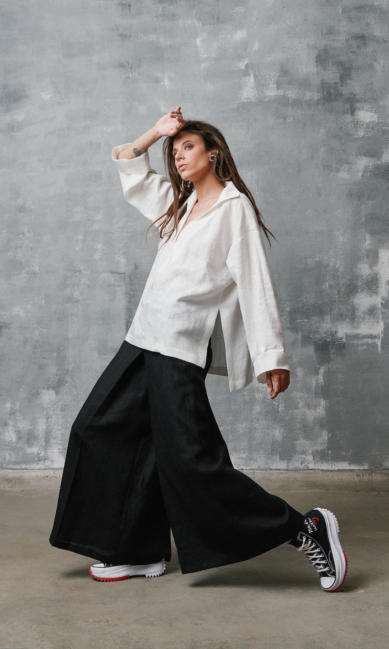 May include: A woman wearing a white linen shirt and black wide-leg pants. She is wearing white sneakers with red accents and black high-top sneakers with red accents.