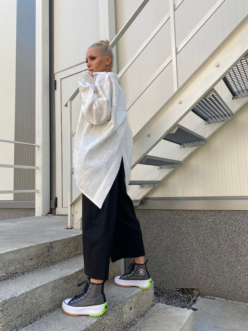 May include: A person wearing a white long-sleeve shirt and black pants. The person is wearing gray high-top sneakers with white soles and neon green accents. The sneakers have a star logo on the side. The person is standing on concrete steps next to a metal staircase.