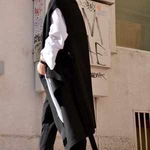 May include: A woman wearing a black sleeveless coat over a white shirt and black pants. She is wearing white sneakers with a heart pattern.