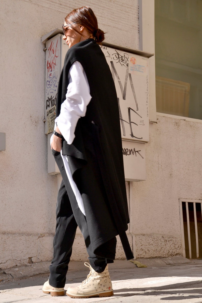 May include: A woman wearing a black sleeveless coat over a white shirt and black pants. She is wearing white sneakers with a heart pattern.
