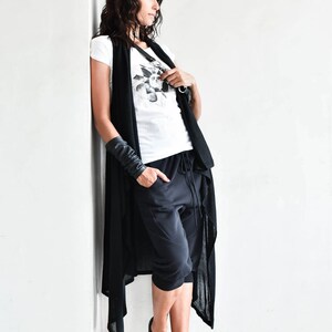 May include: A woman wearing a black long cardigan with a white t-shirt and black pants. She is wearing black leather bracelets and white sneakers.