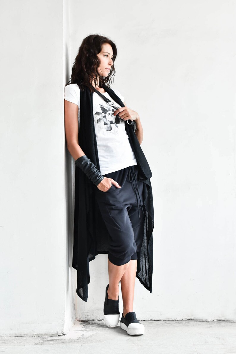 May include: A woman wearing a black long cardigan with a white t-shirt and black pants. She is wearing black leather bracelets and white sneakers.