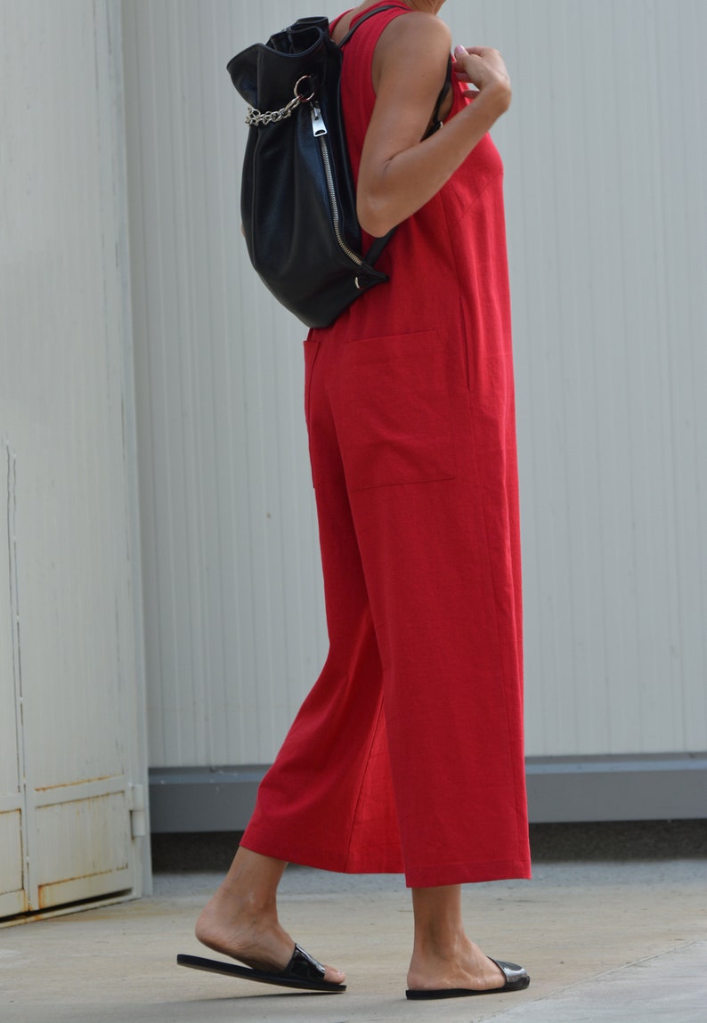 May include: A woman wearing a red sleeveless jumpsuit with a black leather backpack. She is wearing black sandals and walking on a sidewalk.