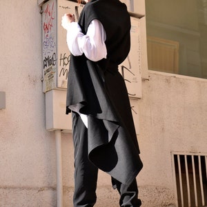 May include: A woman wearing a black sleeveless long vest with a white shirt underneath. She is wearing black pants and white sneakers. The vest has a long, flowing back.