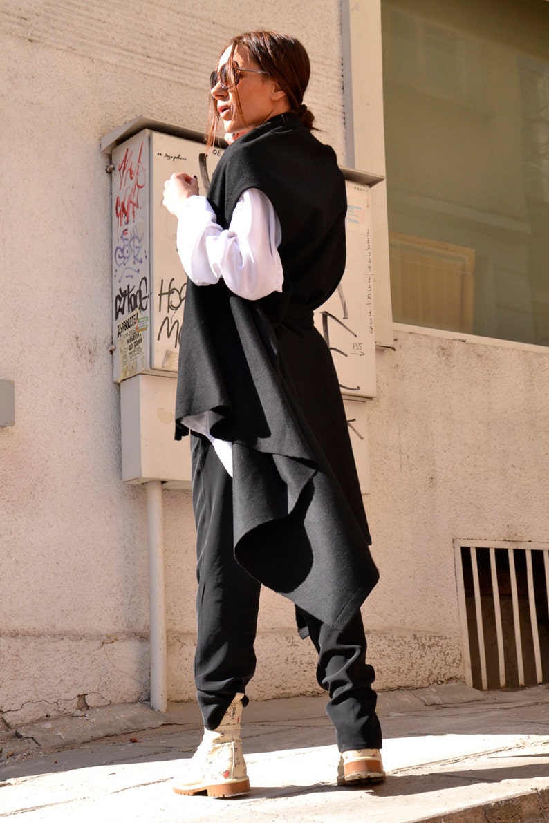 May include: A woman wearing a black sleeveless long vest with a white shirt underneath. She is wearing black pants and white sneakers. The vest has a long, flowing back.