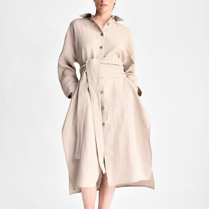 May include: A beige linen shirt dress with a long sleeve and a tie belt. The dress has a button-up front and a relaxed fit.