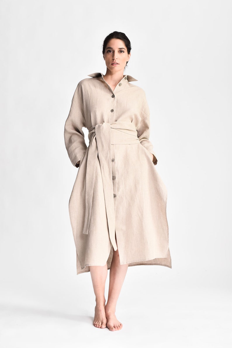 May include: A beige linen shirt dress with a long sleeve and a tie belt. The dress has a button-up front and a relaxed fit.