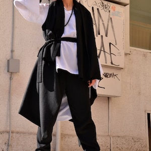 May include: A woman wearing a black long vest with a white shirt underneath, black pants, and beige boots. She is wearing sunglasses and has her hair pulled back.