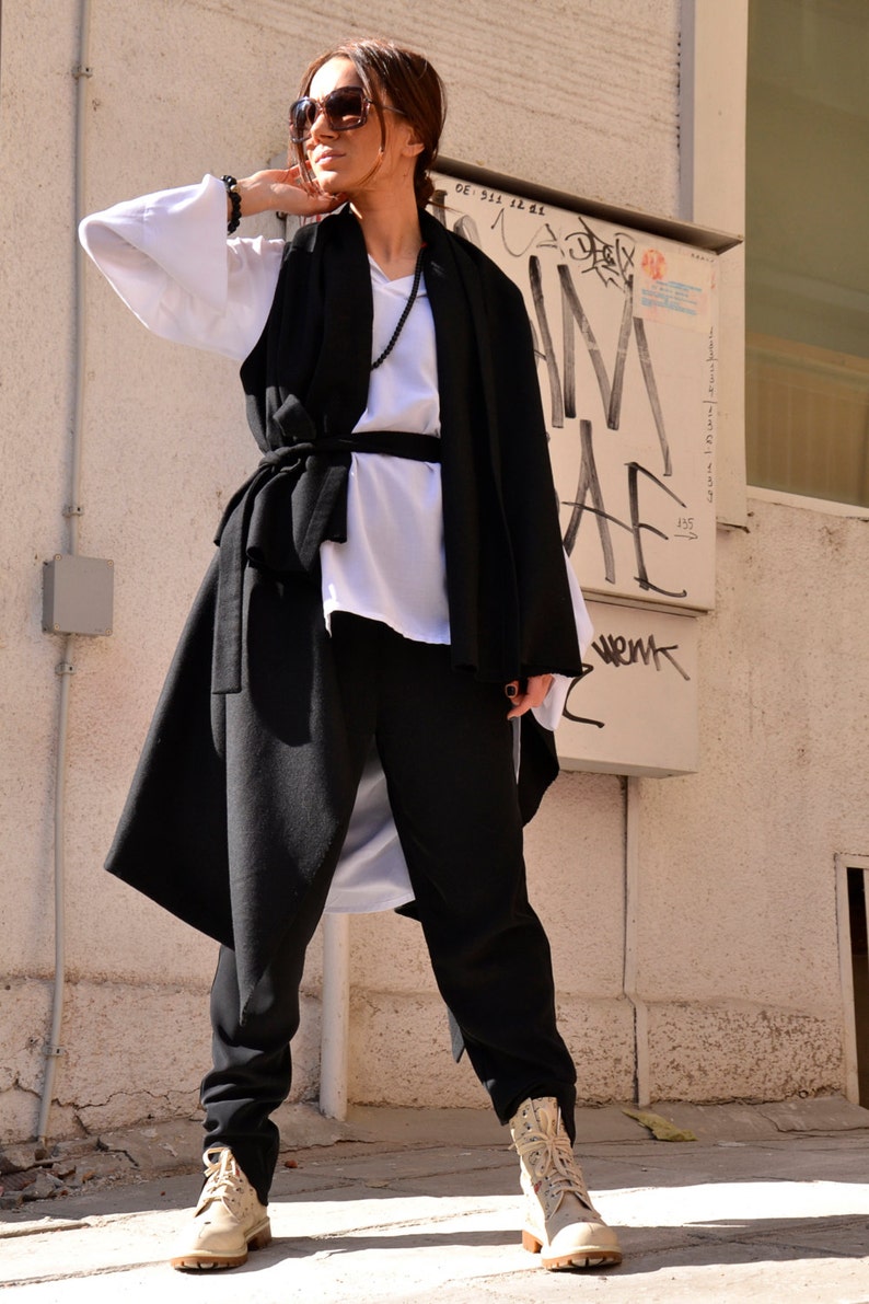 May include: A woman wearing a black long vest with a white shirt underneath, black pants, and beige boots. She is wearing sunglasses and has her hair pulled back.